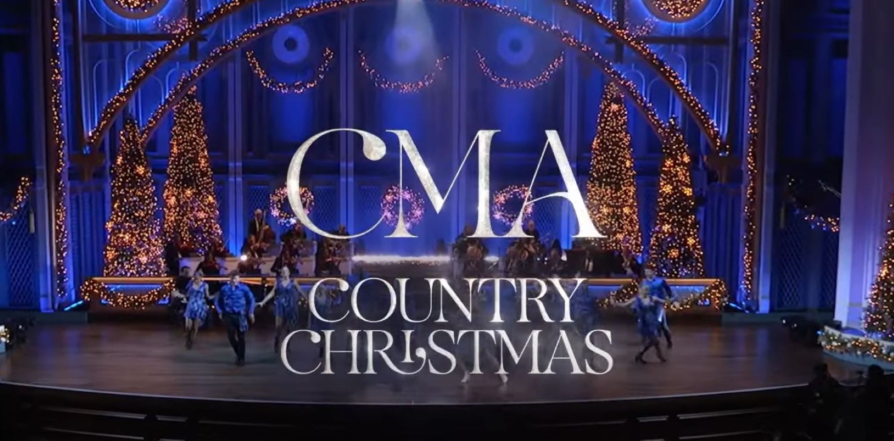 A stage with a blue backdrop and multiple warmly lit Christmas trees. The text overlay reads "CMA Country Christmas."