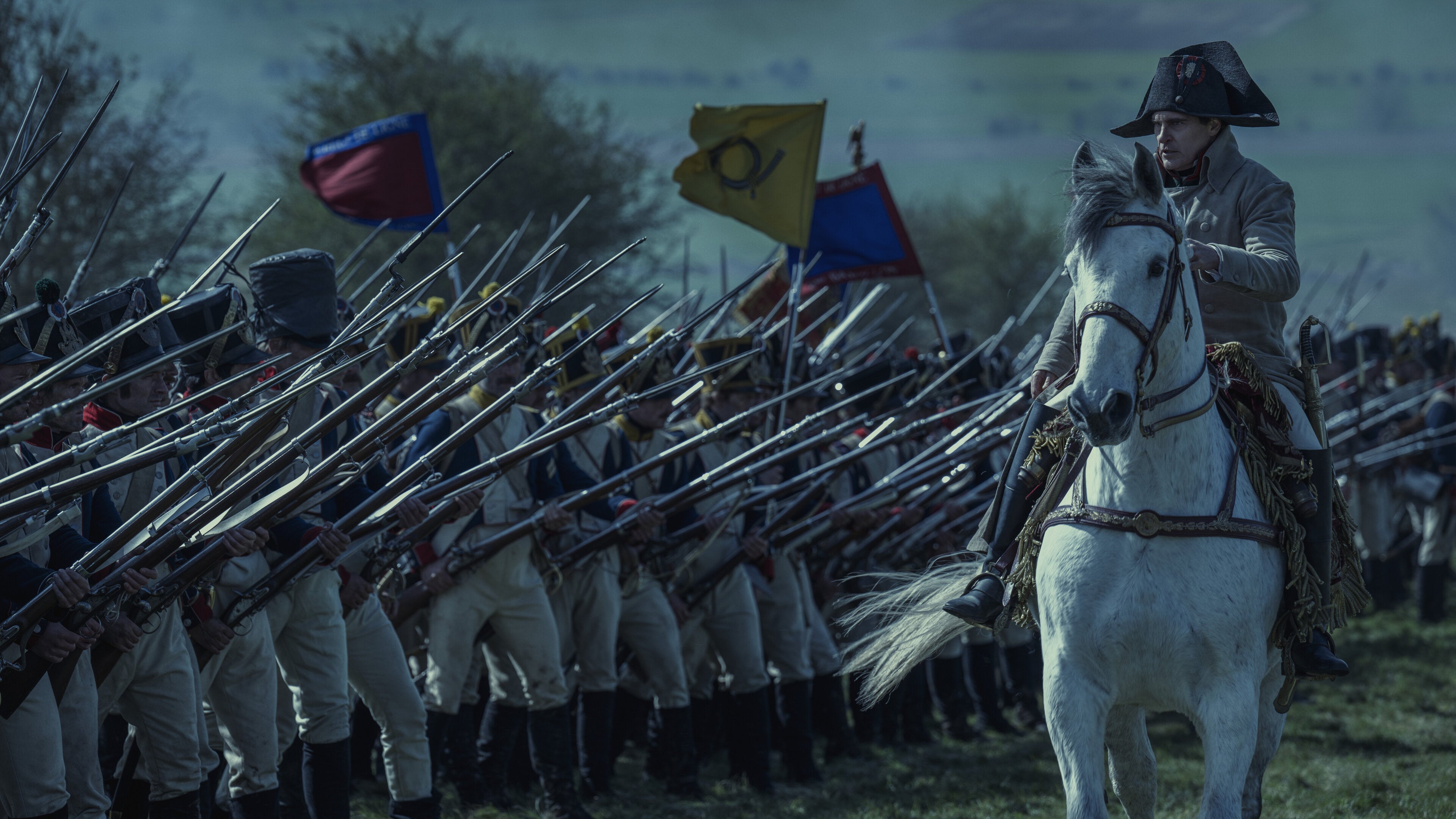 A still from Ridley Scott's "Napoleon" of Joaquin Phoenix dressed in military costume as Napoleon Bonaparte on horseback. He is commanding a large battalion of infantrymen with their bayonets in guard position.