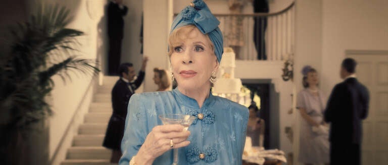 Carol Burnett dressed in a blue silk period jacket and turban holding a martini in a promotional still for the Apple TV+ series "Palm Royale"