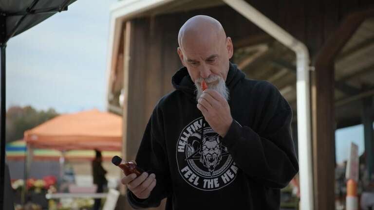 A man in a black sweatshirt with white writing that says "Hellfire Hot Sauce" on the first line with the company logo in the center and "Feel the Heat" on the second line. He is holding a pepper he is sniffing in his left hand and a bottle of hot sauce in his right hand.