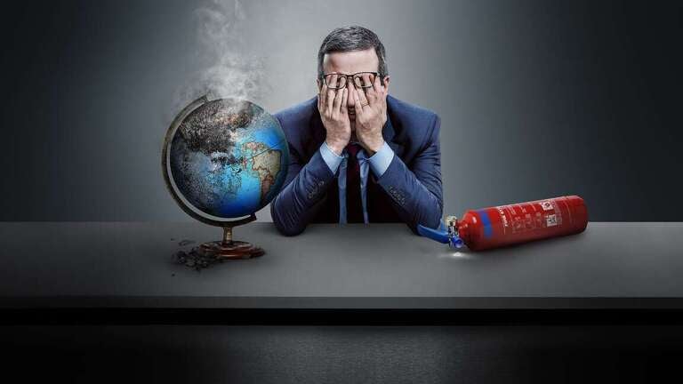 John Oliver sits in a blue suit at a desk covering his eyes with his hands under his glasses. To his right sits a smoking globe, and to his left a recently used fire extinguisher laying on its side