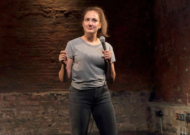 Jacqueline Novak, in black jeans and a gray t-shirt and holding a microphone, performs her stand-up show "Get On Your Knees"
